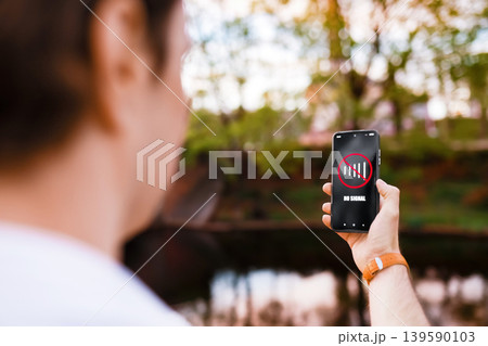 A man holds a smartphone displaying a "No Signal" icon while outdoors in a park. This image illustrates a loss of cellular network, poor connection, or an internet outage. A man holds a smartphone displaying a "No Signal" icon while outdoors in a park. This image illustrates a loss of cellular network, poor connection, or an internet outage. 139590103