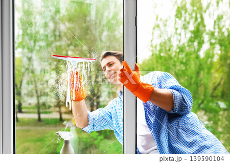 A man in orange gloves uses a squeegee and soapy water to wash a window. This bright image captures a spring cleaning routine, highlighting home maintenance and domestic chores. A man in orange gloves uses a squeegee and soapy water to wash a window. This bright image captures a spring cleaning routine, highlighting home maintenance and domestic chores. 139590104