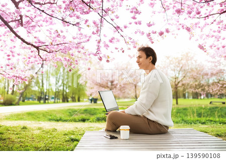 A man works on a laptop under blooming pink cherry blossoms in a park. He sits cross-legged on a wooden platform, enjoying a peaceful remote work lifestyle during springtime. A man works on a laptop under blooming pink cherry blossoms in a park. He sits cross-legged on a wooden platform, enjoying a peaceful remote work lifestyle during springtime. 139590108