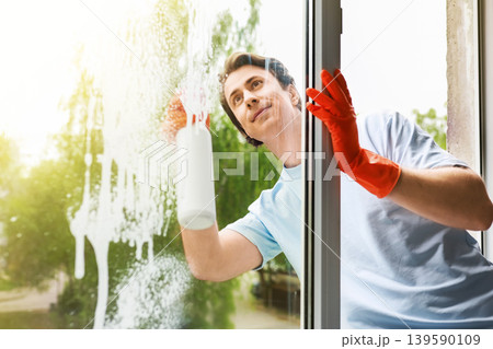 A man wearing red rubber gloves cleans a window with spray detergent and a cloth. This bright image captures a spring cleaning moment, showing home maintenance and chores. 139590109