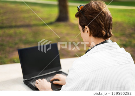 Over-the-shoulder view of a man typing on a laptop while working outdoors in a sunny park. A person sits at a table, demonstrating a remote work and digital nomad lifestyle. 139590282