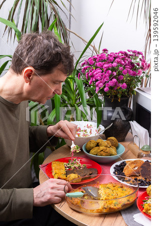 A man sits at a round table and eats various dishes made from homemade recipes. He is surrounded by plants and a bouquet of flowers in a sunny kitchen during breakfast time A man sits at a round table and eats various dishes made from homemade recipes. He is surrounded by plants and a bouquet of flowers in a sunny kitchen during breakfast time 139592046