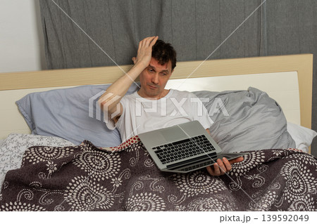 A man stretches while sitting up in bed with a laptop in front of him. It is morning and he has just woken up. The bed appears messy with blankets and sheets around him A man stretches while sitting up in bed with a laptop in front of him. It is morning and he has just woken up. The bed appears messy with blankets and sheets around him 139592049