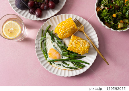 A healthy breakfast is served on a pink table. There are plates with asparagus, a fried egg, corn, and a fresh salad. Grapes and sunflower seeds are on the side A healthy breakfast is served on a pink table. There are plates with asparagus, a fried egg, corn, and a fresh salad. Grapes and sunflower seeds are on the side 139592055