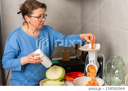 A woman is working in a kitchen to prepare fermented cabbage. She is using a grater and mixing in shredded carrots while measuring salt. The kitchen has different utensils and jars A woman is working in a kitchen to prepare fermented cabbage. She is using a grater and mixing in shredded carrots while measuring salt. The kitchen has different utensils and jars 139592064