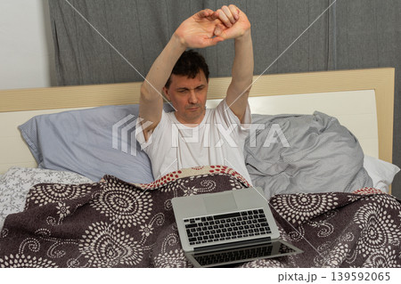 A man stretches while sitting up in bed with a laptop in front of him. It is morning and he has just woken up. The bed appears messy with blankets and sheets around him 139592065
