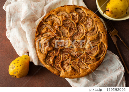 A fresh pear pie is placed on a cloth on a table. Nearby, a bowl holds ripe pears and a small vase has flowers. The setting looks warm and inviting for sharing a treat 139592093