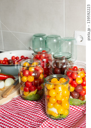 In a kitchen, jars filled with canned tomatoes sit on a table beside fresh vegetables. A mixing bowl holds whole tomatoes as preparation for winter storage continues 139592223
