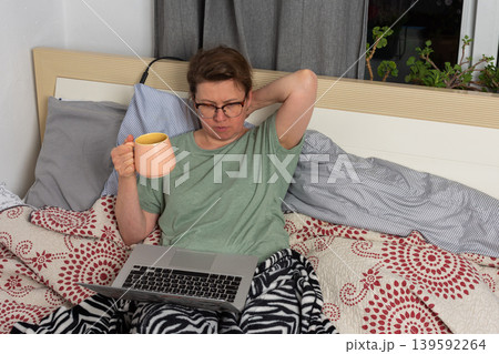 A 50-year-old woman sits on her bed using a laptop and holding a warm drink. She appears focused on her screen while surrounded by cozy blankets in her bedroom A 50-year-old woman sits on her bed using a laptop and holding a warm drink. She appears focused on her screen while surrounded by cozy blankets in her bedroom 139592264