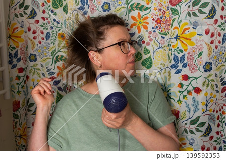 A woman prepares for the day in her bathroom. She is using a hair dryer to style her hair while standing in front of a vibrant shower curtain. Morning light fills the room 139592353