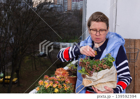 A 50-year-old woman tends to her autumn flowers on a small balcony. She sorts plants from a bag while enjoying her time outdoors in a city environment A 50-year-old woman tends to her autumn flowers on a small balcony. She sorts plants from a bag while enjoying her time outdoors in a city environment 139592394