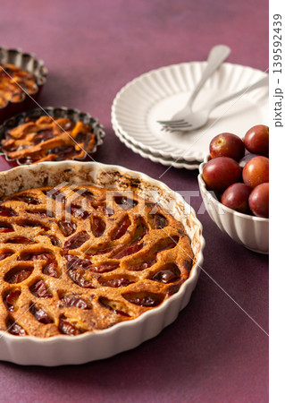 A plum pie is placed on a table with thinly sliced plums visible on top. Bowls of fresh plums are nearby with empty plates waiting for dessert 139592439