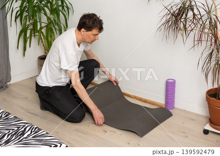 A 53-year-old man is kneeling on the floor in his home setting. He is laying out an exercise mat for his workout. Nearby, there are green plants and a purple foam roller A 53-year-old man is kneeling on the floor in his home setting. He is laying out an exercise mat for his workout. Nearby, there are green plants and a purple foam roller 139592479