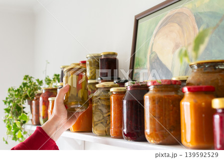 A person holds a jar of canned vegetables while checking the shelf filled with various jars of preserved food. This activity takes place during winter preparations for storage 139592529