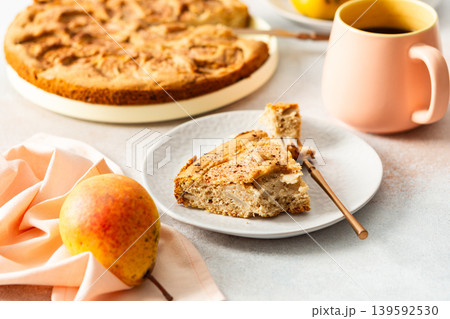 A slice of pear pie sits on a white plate with a fork. A warm coffee cup is next to it. Fresh pears are placed on the table, adding to the scene 139592530