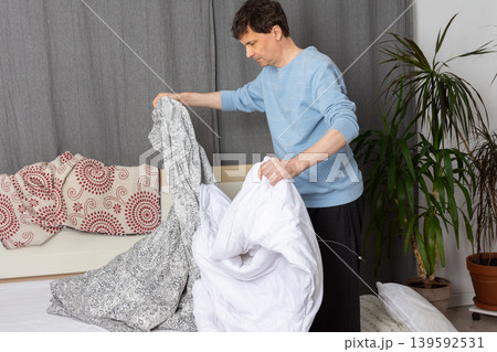 A 53-year-old man is in his bedroom changing bed linen. He holds a patterned bed cover and a white duvet as he prepares to make the bed clean and tidy 139592531