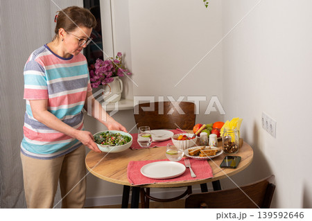 A 53-year-old woman sets the table for dinner. She carries a stack of plates and a glass of water. Fresh fruits and vegetables are arranged on the table, ready for the meal 139592646