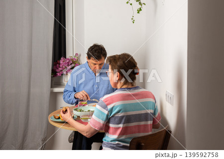A married couple is having dinner together at home. They sit at a small table near a window with a bowl of salad and other food. The light is warm and cozy in the evening A married couple is having dinner together at home. They sit at a small table near a window with a bowl of salad and other food. The light is warm and cozy in the evening 139592758