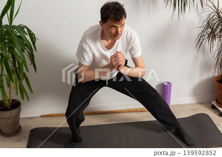 A 53-year-old man practices exercises at home on a yoga mat. He focuses on his strength and flexibility while avoiding distractions from the surroundings. Plants stand around him A 53-year-old man practices exercises at home on a yoga mat. He focuses on his strength and flexibility while avoiding distractions from the surroundings. Plants stand around him 139592852