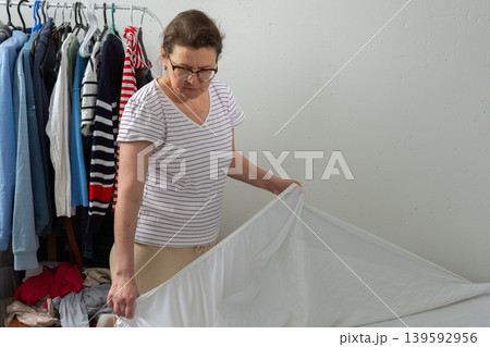 A 53-year-old woman is working in her bedroom. She is changing the bed linen and placing new sheets on the bed. Clothes are seen nearby in the room A 53-year-old woman is working in her bedroom. She is changing the bed linen and placing new sheets on the bed. Clothes are seen nearby in the room 139592956