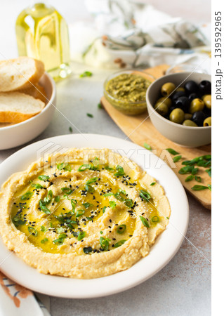 A plate of smooth, creamy hummus drizzled with olive oil sits on a dining table. It is accompanied by bowls of olives, pickles, and fresh bread ready to enjoy A plate of smooth, creamy hummus drizzled with olive oil sits on a dining table. It is accompanied by bowls of olives, pickles, and fresh bread ready to enjoy 139592965