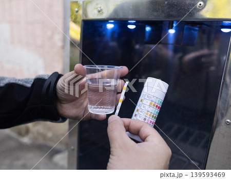 Man checking water quality at a public vending machine using a test strip. Analysis of purified drinking water in a plastic cup. Safety, pH, and hardness control of urban water supply. 139593469