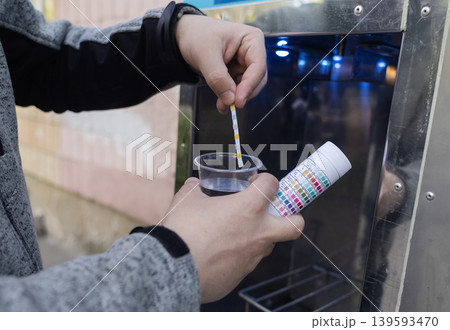 Man checking water quality at a public vending machine using a test strip. Analysis of purified drinking water in a plastic cup. Safety, pH, and hardness control of urban water supply. 139593470