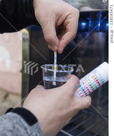 Man checking water quality at a public vending machine using a test strip. Analysis of purified drinking water in a plastic cup. Safety, pH, and hardness control of urban water supply. 139593471