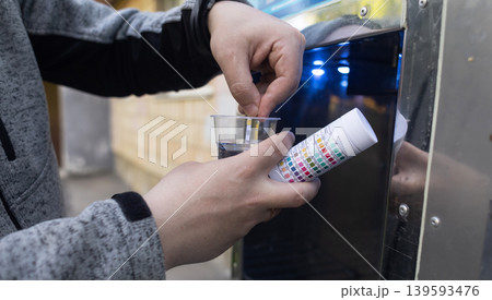 Man checking water quality at a public vending machine using a test strip. Analysis of purified drinking water in a plastic cup. Safety, pH, and hardness control of urban water supply. 139593476