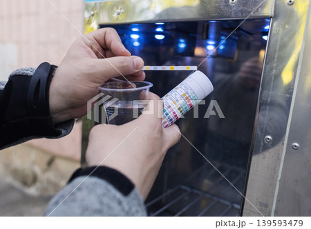 Man checking water quality at a public vending machine using a test strip. Analysis of purified drinking water in a plastic cup. Safety, pH, and hardness control of urban water supply. 139593479