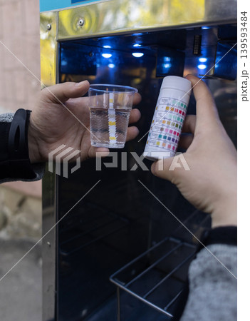 Man checking water quality at a public vending machine using a test strip. Analysis of purified drinking water in a plastic cup. Safety, pH, and hardness control of urban water supply. 139593484