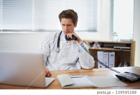 Laptop, phone call and man doctor in his office for telehealth consultation at hospital. Confused, professional and young male healthcare worker on mobile discussion with landline in medical clinic 139595289