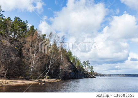 Calm lake shoreline with rocky cliffs and leaning birch trees, pine forest backdrop Calm lake shoreline with rocky cliffs and leaning birch trees, pine forest backdrop 139595553