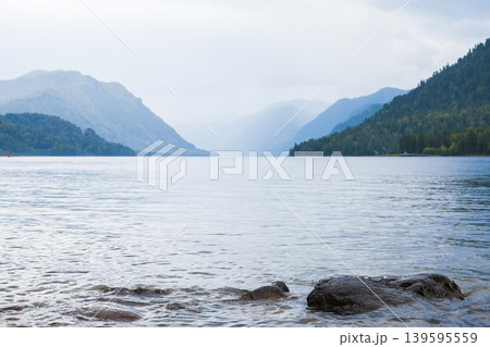 A tranquil mountain lake extends toward misty horizons, framed by forested slopes 139595559