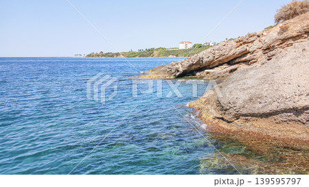 Coastal rocky shoreline with clear blue water and distant buildings Coastal rocky shoreline with clear blue water and distant buildings 139595797