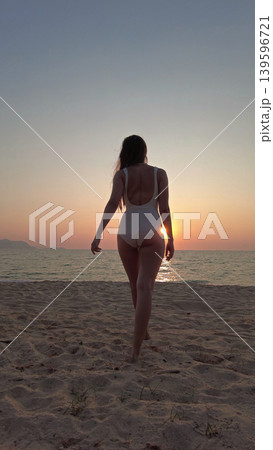 Woman in white swimsuit walking on beach to sunset over sea 139596721