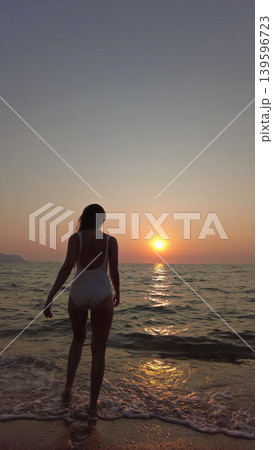 Woman in white swimsuit walking on beach to sunset over sea 139596723