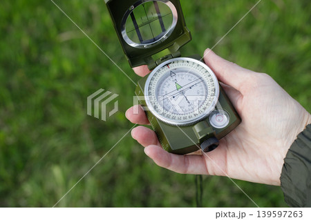 Woman's hand holds an open compass against a background of green grass. Woman navigates her surroundings using the compass. Woman's hand holds an open compass against a background of green grass. Woman navigates her surroundings using the compass. 139597263