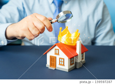A man examines a house with a golden crown under a magnifying glass. Evaluating premium housing. Desirable property qualities and real estate ranking. 139597287