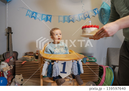 Baby boy sits in high chair looking surprised while father holds a birthday cake with candles. Colorful decorations fill the living room. Family gathers to celebrate first birthday occasion. Baby boy sits in high chair looking surprised while father holds a birthday cake with candles. Colorful decorations fill the living room. Family gathers to celebrate first birthday occasion. 139597640