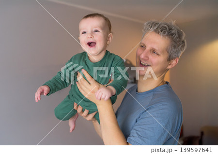 Father and toddler son having fun. Young father lifts his joyful baby in a cozy room. Baby laughing, showing delight. They enjoy their time together at home. Dad playing with baby, happy childhood. 139597641