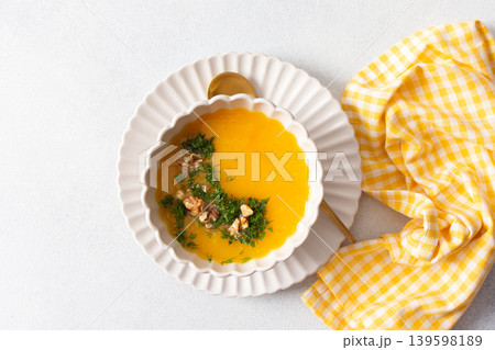 A bowl of pumpkin cream soup sits on a plate. The soup contains nuts and herbs. A yellow checkered napkin lies nearby. This meal can be enjoyed at breakfast or lunch A bowl of pumpkin cream soup sits on a plate. The soup contains nuts and herbs. A yellow checkered napkin lies nearby. This meal can be enjoyed at breakfast or lunch 139598189