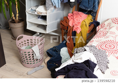 Clothes are scattered on the floor and inside a basket in a room. A chair holds additional clothes. The scene shows items ready for washing after daily use and activities 139598210