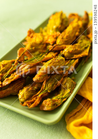 Delicious deep-fried zucchini flowers are neatly arranged on a green platter, showcasing their crispy texture. A bright yellow napkin adds warmth to the scene 139598248
