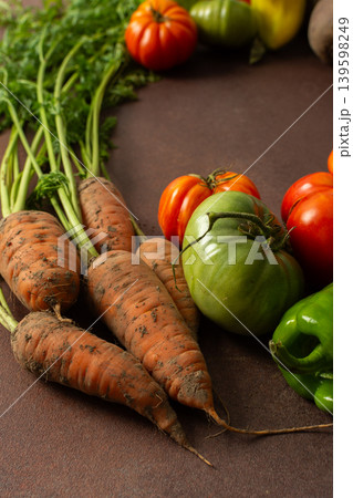 Fresh carrots sit next to red and green tomatoes along with bell peppers on a brown surface. The scene shows a variety of vegetables gathered for cooking or selling 139598249
