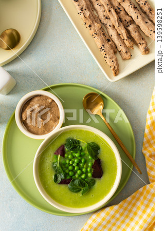 A creamy soup made from broccoli and green peas is served alongside liver pate and grissini sticks on a simple table setting. Fresh ingredients are arranged neatly for a healthy meal 139598251