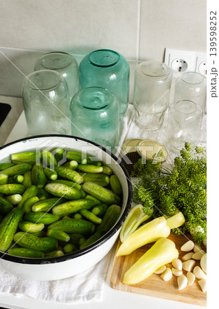 Cucumbers are soaking in a bowl while garlic and cucumber slices are on a cutting board. Several empty jars sit nearby on a clean countertop. It is a preparation activity for winter storage Cucumbers are soaking in a bowl while garlic and cucumber slices are on a cutting board. Several empty jars sit nearby on a clean countertop. It is a preparation activity for winter storage 139598252