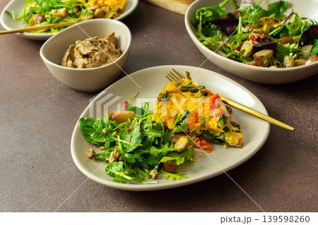 A plate features baked eggs with vegetables alongside a fresh arugula salad. Small bowls hold liver pate and baked pear pieces. This meal is set on a neutral table 139598260