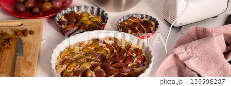 Cook is preparing to make plum pie in a home kitchen. Ingredients like plums, flour, sugar, and butter are on the counter. Tools for mixing and baking are nearby, banner 139598287