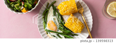 A healthy breakfast is served on a pink table. There are plates with asparagus, a fried egg, corn, and a fresh salad. Grapes and sunflower seeds are on the side, banner 139598313